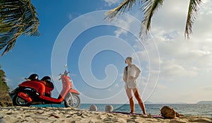 Scooter road trip. Lovely couple on red motorbike in white clothes on sand beach. People walking near the tropical palm