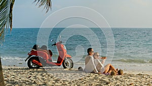 Scooter road trip. Lovely couple on red motorbike in white clothes on sand beach. People walking near the tropical palm