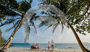 Scooter road trip. Lovely couple on red motorbike in white clothes on sand beach. People walking near the tropical palm