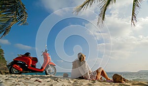 Scooter road trip. Lovely couple on red motorbike in white clothes on sand beach. People walking near the tropical palm