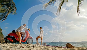 Scooter road trip. Lovely couple on red motorbike in white clothes on sand beach. People walking near the tropical palm