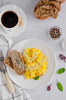 Scrambled eggs cooking from organic fresh eggs on a white plate with rye bread on a light background with a cup of coffee.