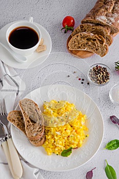 Scrambled eggs cooking from organic fresh eggs on a white plate with rye bread on a light background with a cup of coffee.