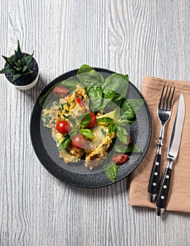 Scrambled eggs omelette portion for breakfast decorated with basil leaves and cherry tomatoes on a black porcelain plate. Knife