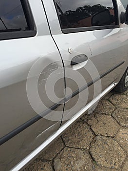 Scratched and dented side of a silver gray car right next to the door handle, with wall paint marks in need of a body shop and