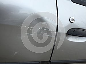 Scratched and dented side of a silver gray car right next to the door handle, with wall paint marks.