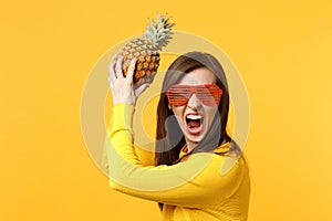 Screaming crazy young woman in funny glasses holding fresh ripe pineapple fruit isolated on yellow orange wall