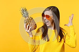 Screaming crazy young woman in heart glasses spreading hands holding fresh ripe pineapple fruit isolated on yellow