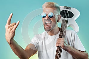 Screaming hipster man holding white bass guitar and show rock gesture in neon lights. Rock music concept.