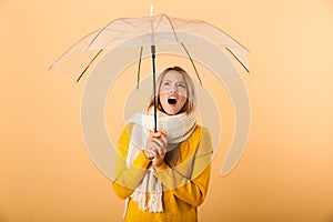 Screaming shocked woman wearing scarf holding umbrella posing over yellow wall background