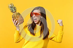 Screaming woman in funny glasses clenching fist like winner holding fresh ripe pineapple fruit isolated on yellow orange