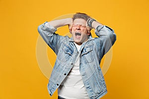 Screaming young man in denim casual clothes keeping eyes closed, putting hands on head isolated on yellow orange