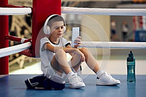 Scrolling social media feed after training. Life portrait of little boy, beginner boxer in sport uniform during workout