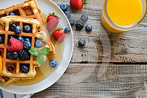 Scrumptious waffles with syrup, summer berries, and orange juice on a rustic wooden table. top view