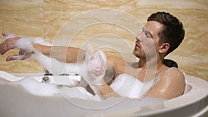 Scrupulous man soaping himself with loofah, taking bath with foam, cleanness