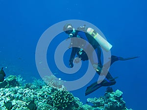 The scuba diver turns to look at the camera. Blue water deep is on the background. The scuba diver is wearing in full equipment: t