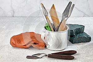Sculpting and modeling tools in aluminum mug surrounded by red clay and green plasticine on light stone table