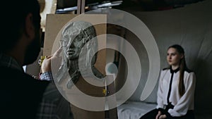 Sculptor creating sculpture of human`s face on canvas while young woman posing him in art studio