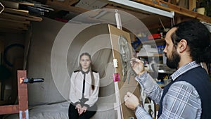 Sculptor creating sculpture of human`s face on canvas while young woman posing to him in art studio