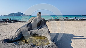 A sculpture of a mermaid made of stone on a sandy beach and a volcanic seashore with people walking on the stones