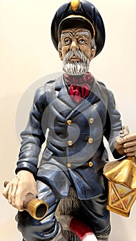 A sculpture of a sailor stands looking forward he has a gray beard mustache and a cap And a spyglass in his hand and a