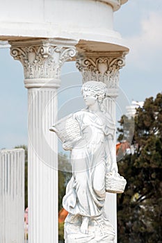 Sculpture of young woman holding basket full of grapes or fruits near corinthian columns in a public park. Replica of