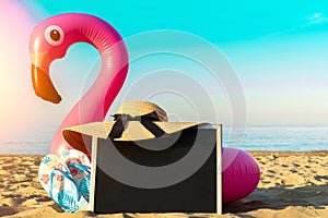 Sea background. Funny pink toy flamingo with blackboard, slippers and hat for text on summer ocean nature beach background in