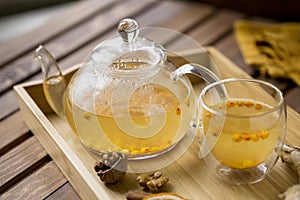 Sea buck thorn tea in glass teapot with glass cup and spoon, orange and walnuts near, at wooden tray on wood background