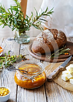 Sea buckthorn jam in a glass jar with butter, fresh berries, leaves and loaf bread