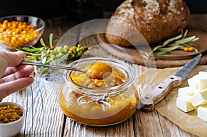 Sea buckthorn jam in a glass jar with spoon an woman hand, sliced butter, fresh berries, leaves and loaf bread