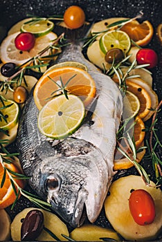 Sea gilt-head bream fish in the baking tray ready to be baked with potatoes, rosemary, lemon, orange, olives, tomatoes, onion and