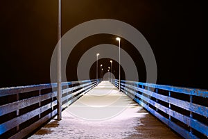 Sea pier covered with snow in night yellow lights from lampposts, mysterious path in darkness