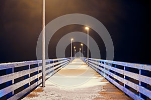 Sea pier covered with snow in night yellow lights from lampposts, mysterious path in darkness