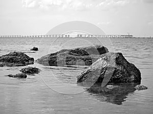 Sea, rocks, bridge in the background. Black and white photography emphasizes the contrast between stone and water.