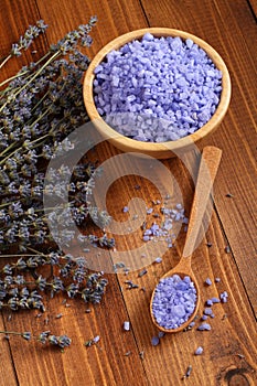 Sea salt with lavender in a bamboo bowl and in a wooden spoon and dried lavender flowers are located on a brown wooden table.