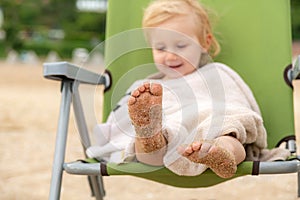 Sea sand on the feet of a child. A little girl covered with a towel sits on a folding chair, her feet are wet in the sand