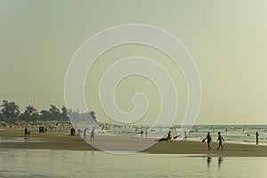 Sea sandy beach in the evening against the backdrop of huts and trees, a lot of people walking to the beach and swimming in the