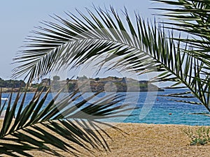 Sea view from the shore towards a fortress in Sithonia peninsula, Halkidiki, with a palm tree leaf close up in front.