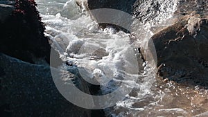 Sea water gushing through a gap between large rocks on the beach. Slow motion from above.