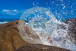 Sea water splashes against the background of the ocean and blue sky. A large ocean wave breaks violently on the rocks