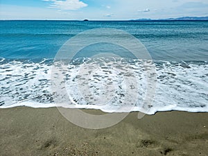 Sea water white foam on wet sand, blue sky. Empty sandy beach, Greek island Cyclades, Greece. Space