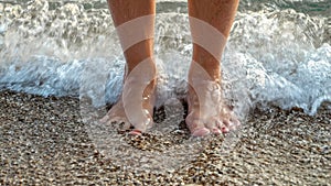 Sea waves rolling and breaking on female feet standing on the ocean beach. Holiday, summer vacation and tourism
