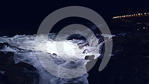 Sea waves splash and crash into rocks with much white foam. Night cinematic shot sea violently splashing over the rocky coast