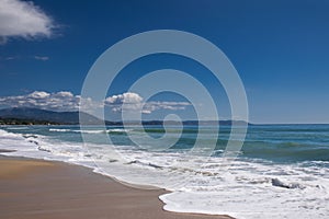 Sea waves and white foam in paradise sandy beach, blue sky, white clouds, sun.