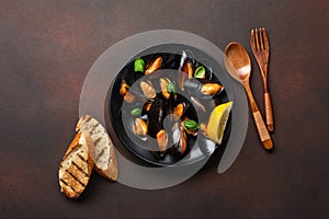 Seafood mussels and basil leaves in a black plate with towel, bread slices, wooden spoon and fork on rusty background