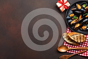 Seafood mussels and basil leaves in a black plate with towel, bread slices, wooden spoon and fork on rusty background