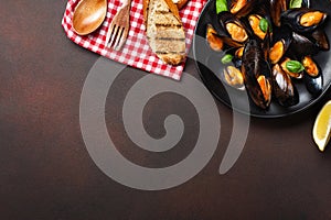 Seafood mussels and basil leaves in a black plate with towel, bread slices, wooden spoon and fork on rusty background