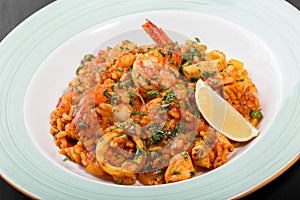 Seafood paella with rice, tomato sauce, langoustine, mussels, squid, shrimp and greens on plate on black wooden background.