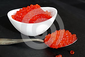 Red caviar in a white plate with a spoon lies on a black background