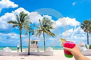 Seafront beach promenade with hand holding cocktail and palm trees on background on sunny day in Fort Lauderdale in